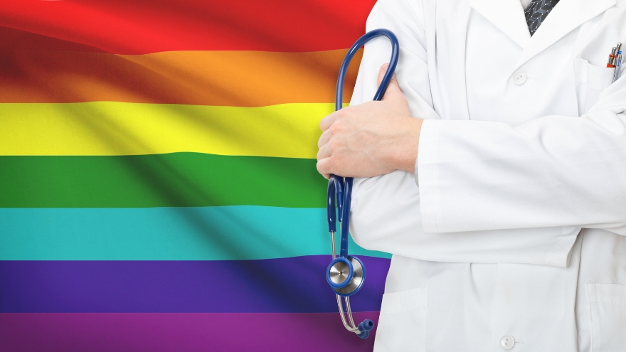 A doctor stands in front of an LGBTQIA Pride flag holding a stethoscope.